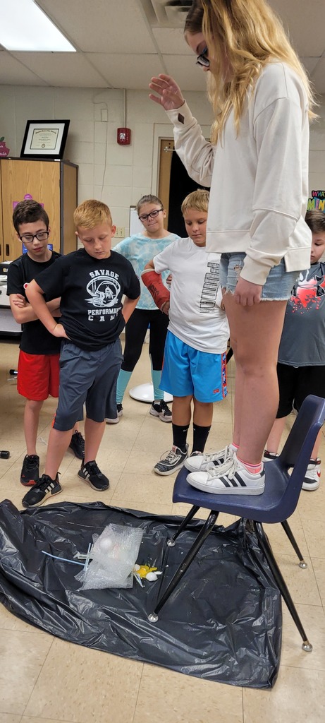 students watch as another student drops an egg while standing on a chair 