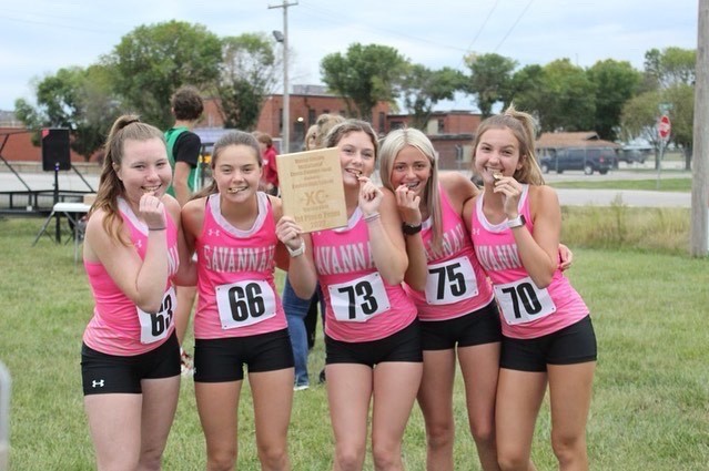 5 girls in pink xc uniforms pose for a picture with their gold medals and a plaque for taking 1st place at the benton invitational cross country meet. 