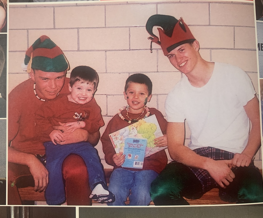 two high school aged boys dressed as elves sit with two elementary aged boys and read picture books