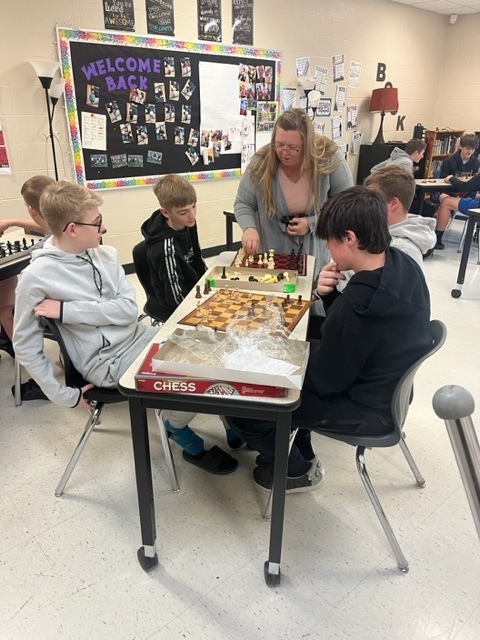 four students learn to play chess while a teacher watches over them 