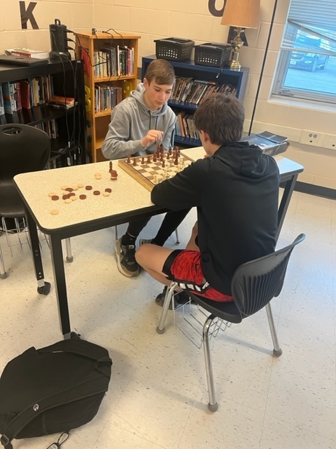 two students sit across a table from each other and play chess 